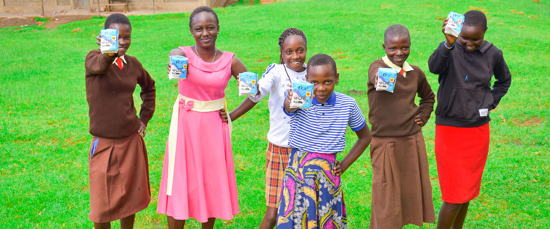 Six smiling girls stand on grass, each holding up a cup of yogurt. Wearing brightly colored dresses, they appear joyful and proud—celebrating support from the Collaborative Fund in raising awareness about period poverty outdoors together.