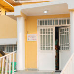 Entrance to a yellow building with white doors, columns, and a ramp. A sign near the entrance indicates comprehensive abortion care is offered here. Laundry dries on the left, and shrubs grow close to the ramp.