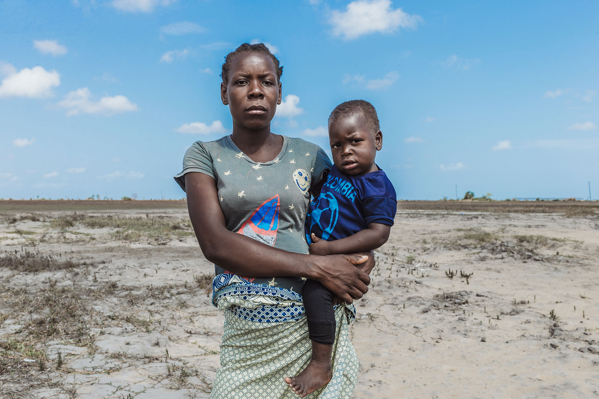 A woman stands outdoors on dry, barren land, holding a young child in her arms. Both look directly at the camera with serious expressions. The sky is blue with a few scattered clouds.