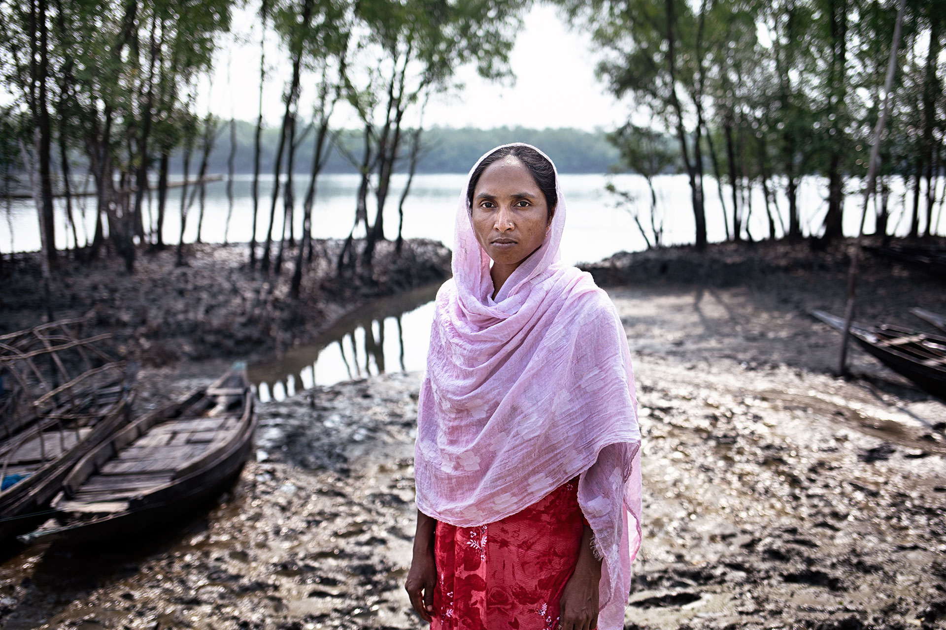 A woman wearing a pink scarf and red dress stands on muddy ground near wooden boats and trees by a riverbank with water and forest in the background.
