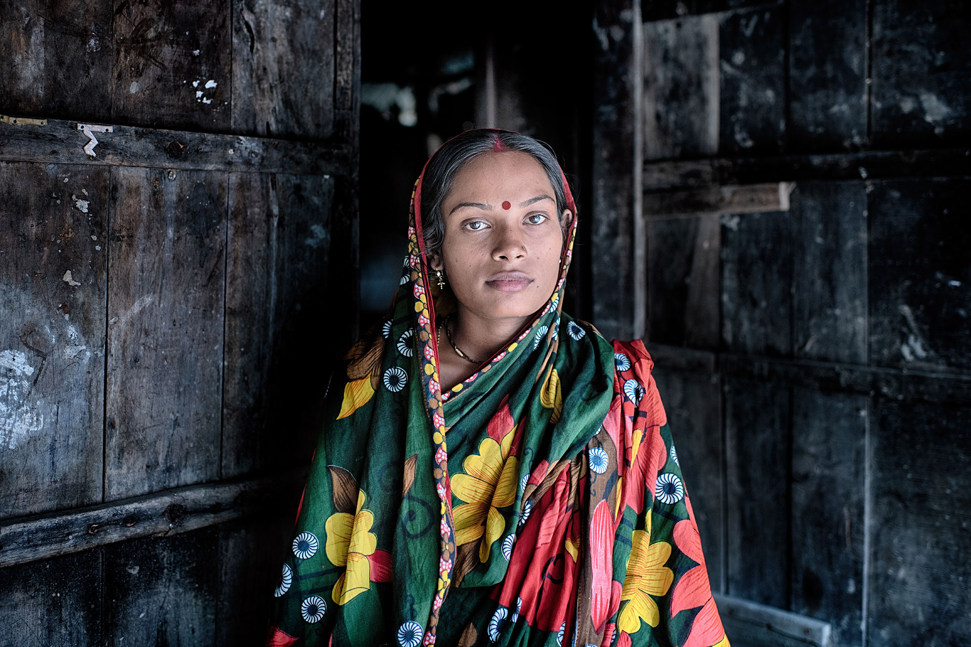 A woman in a colorful floral saree stands in a doorway, looking directly at the camera. The background is made of dark, weathered wooden planks, creating a rustic setting.