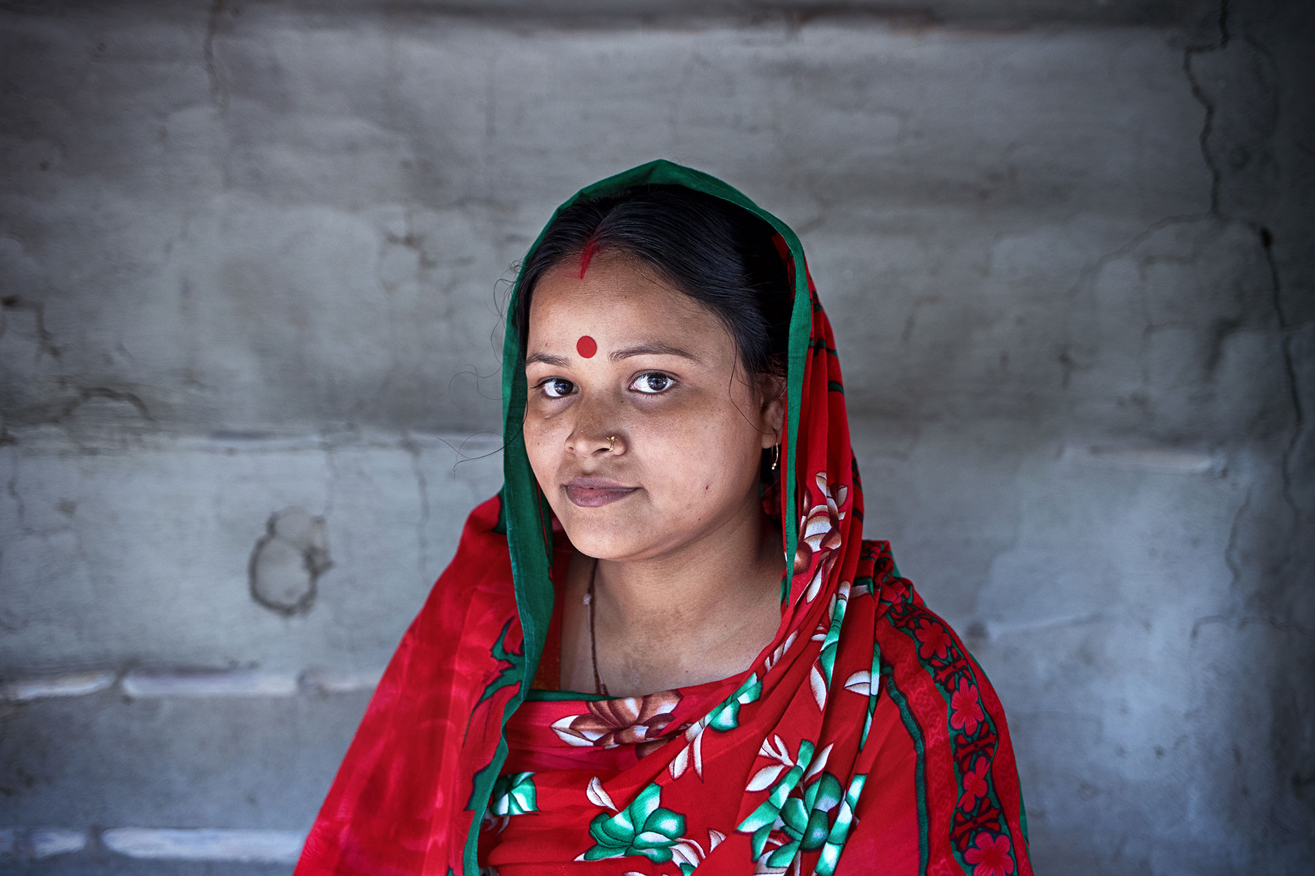 A woman wearing a red and green floral sari with a red bindi stands in front of a textured gray wall, looking calmly at the camera.