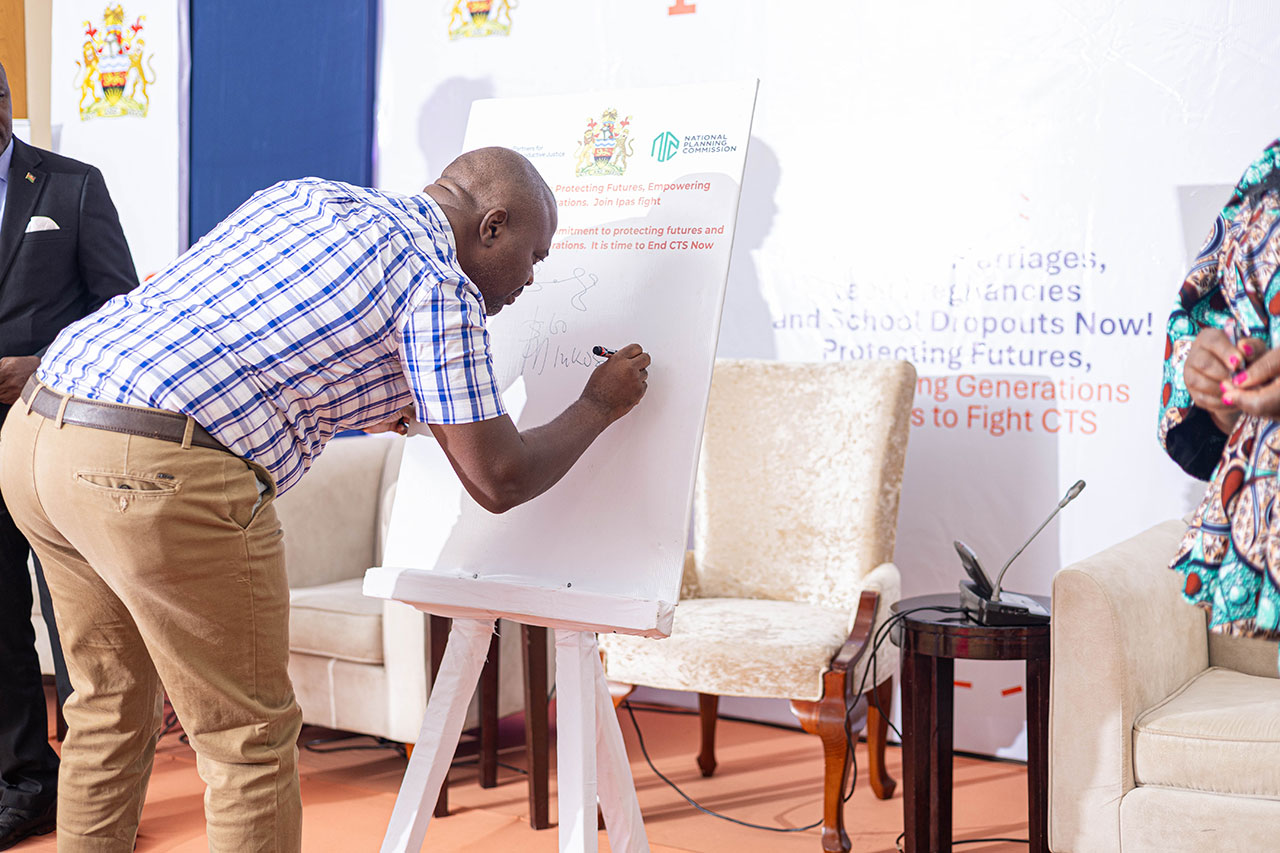 Senior Chief Mabulawo, signing the commitment for CTS support. A man in a checked shirt signs a document on an easel in a well-lit room. There are two chairs in the background and a banner with colorful text and logos.