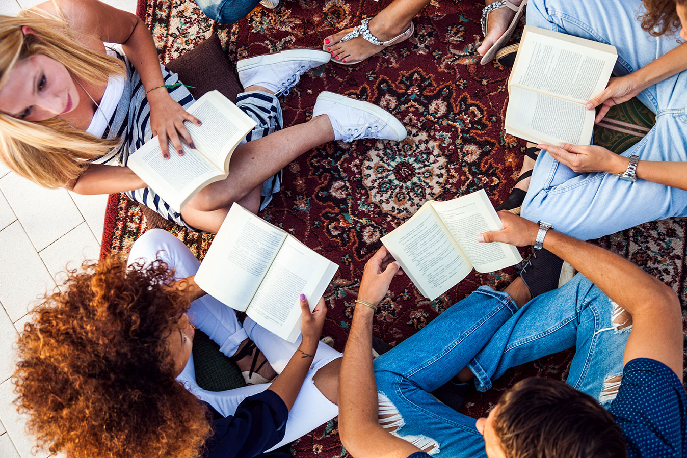 Reading with friends A group of people sitting on a patterned rug, forming a circle. They are reading books, with crisp pages visible. The individuals appear engaged, some looking at the open books, dressed casually with a variety of styles and colors in clothing.