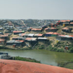 A wide view of a densely crowded hillside refugee camp with rows of makeshift shelters covered in orange and black tarps, surrounding a small pond under a hazy sky highlights the risks, like unsafe abortion in refugee camps, faced by residents.