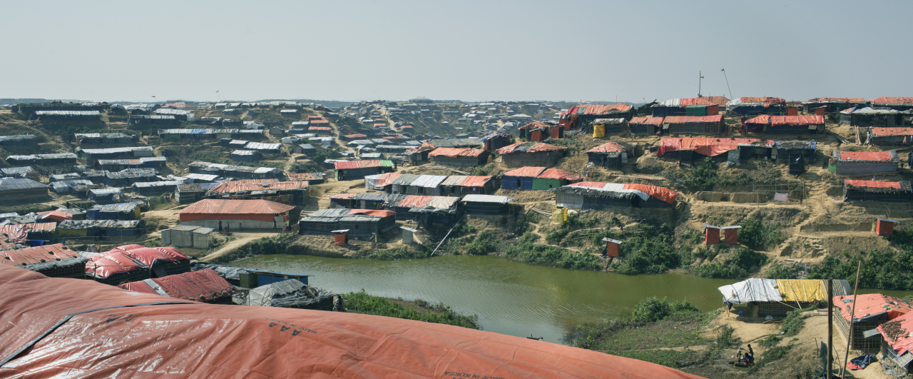 A wide view of a densely crowded hillside refugee camp with rows of makeshift shelters covered in orange and black tarps, surrounding a small pond under a hazy sky highlights the risks, like unsafe abortion in refugee camps, faced by residents.