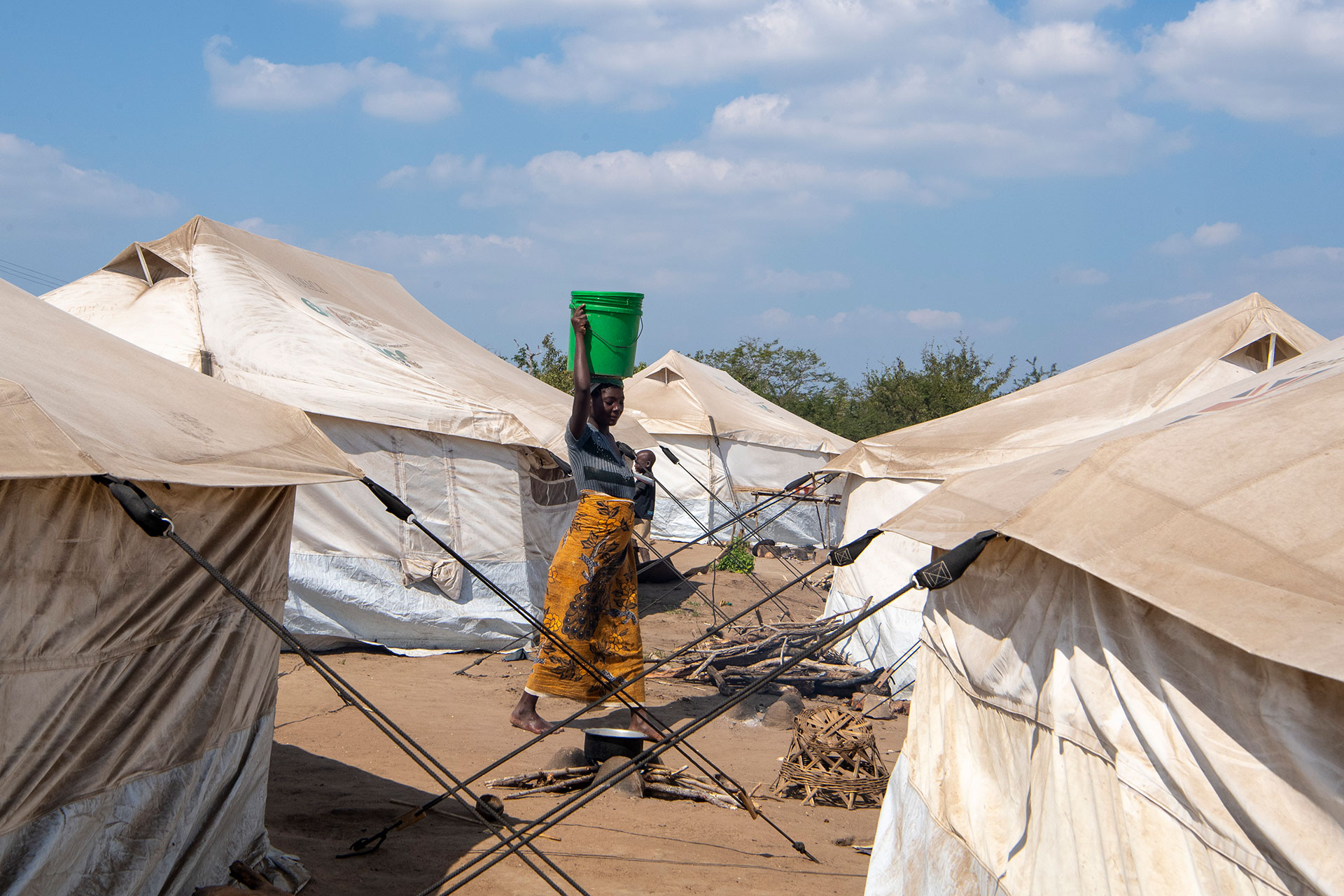 Damage from Cyclones Idai and Kenneth. A woman in a yellow patterned skirt carries a green bucket on her head, walking between white tents in a camp under a blue sky with scattered clouds.