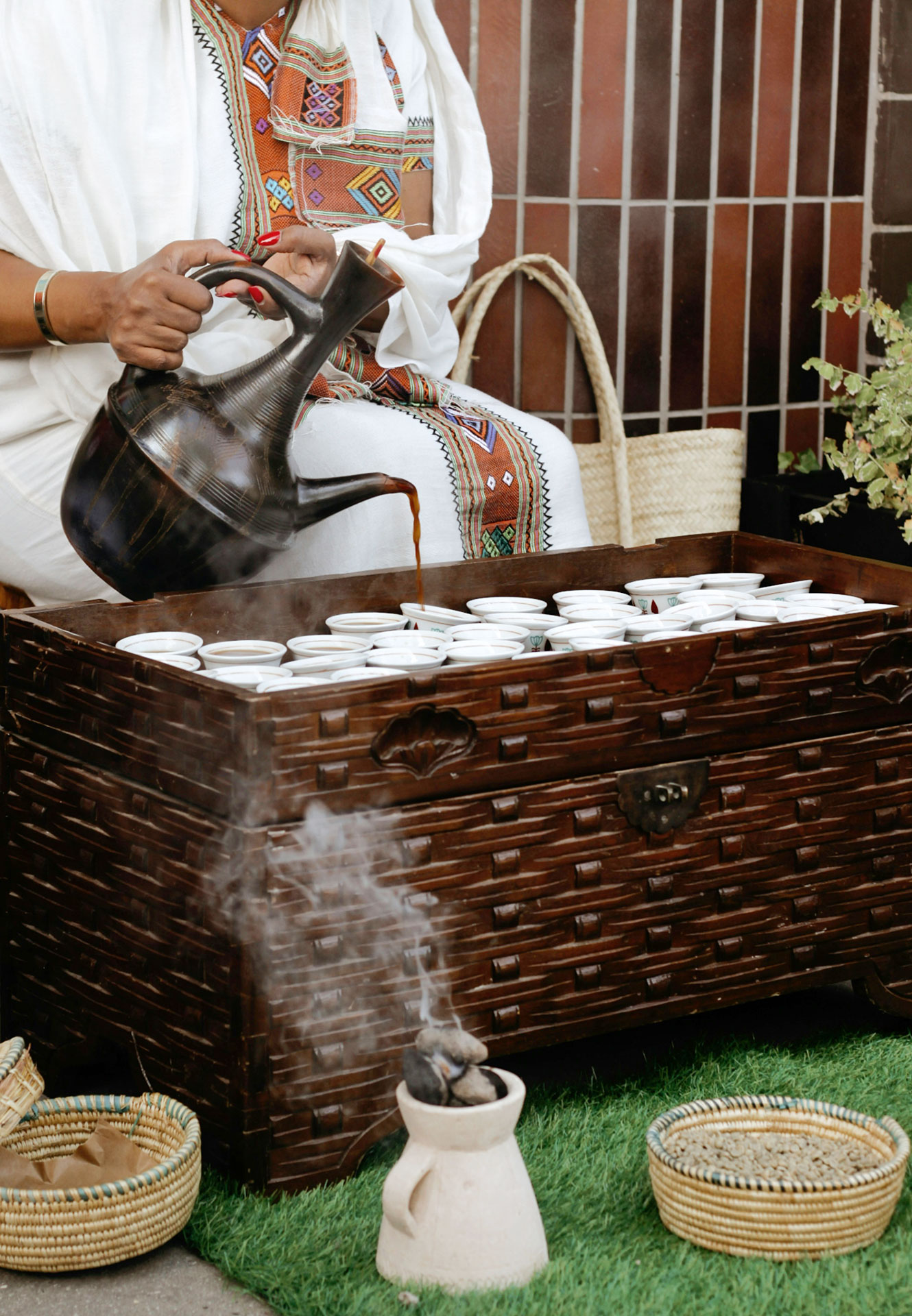 Ethiopian coffee ceremony A person in traditional attire pours coffee from a pot into small cups arranged on a wooden chest, with woven baskets and incense burning on the grass in front.