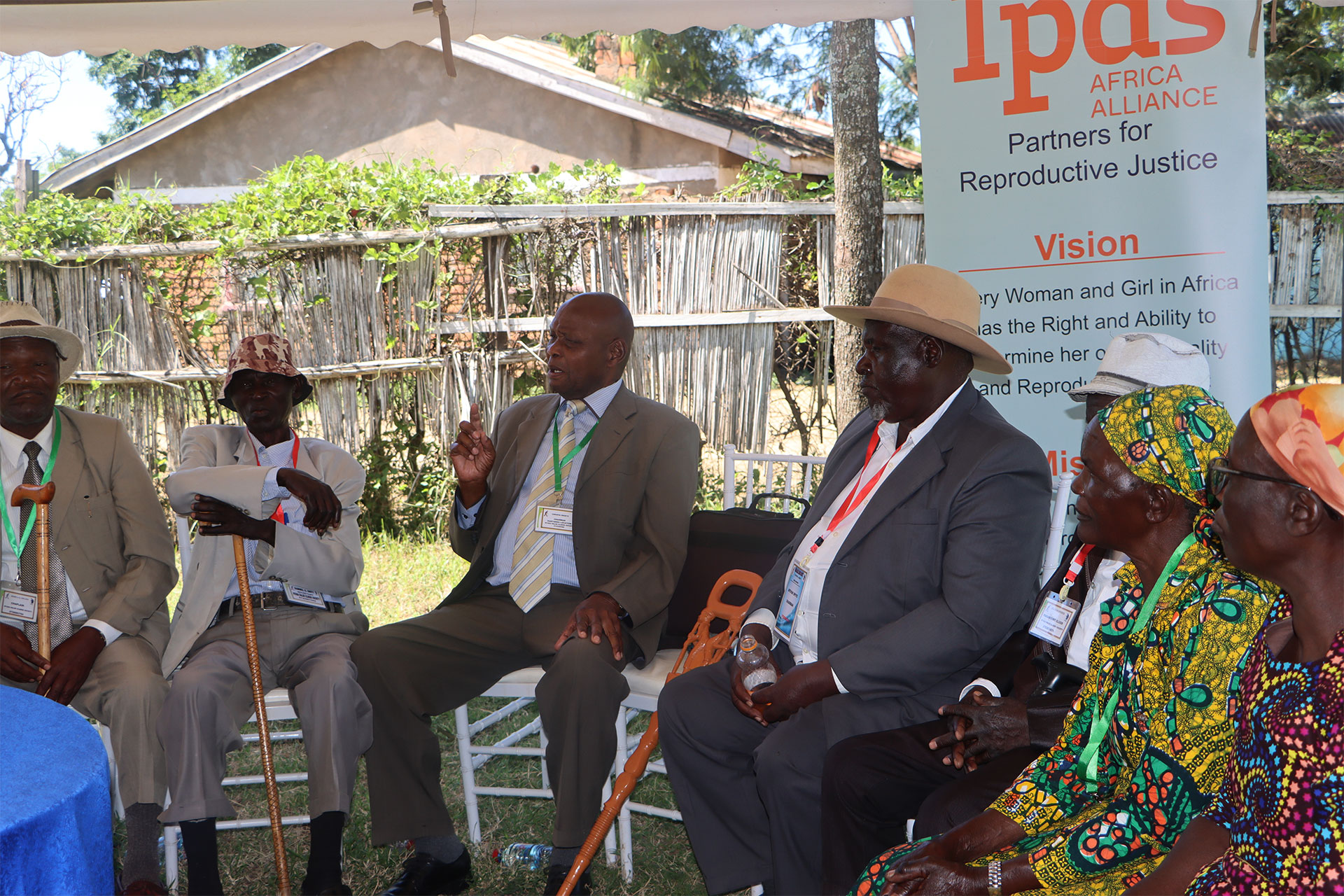 A section of Nyadaralek Elders discussing strategies to engage adolescents in the county A group of men and women in traditional clothing sit in a circle outdoors, engaged in a discussion on Kenya adolescent SRHR and the triple threat. A banner for Ipas Africa Alliance, Partners for Reproductive Justice, is visible in the background.