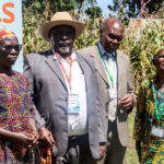 A group of six adults, four women and two men, stand together outdoors in Kenya, smiling. Dressed in colorful clothing with name badges, they gather before a display board and a cornfield to discuss adolescent SRHR and the triple threat.