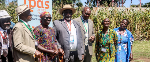 A group of six adults, four women and two men, stand together outdoors in Kenya, smiling. Dressed in colorful clothing with name badges, they gather before a display board and a cornfield to discuss adolescent SRHR and the triple threat.