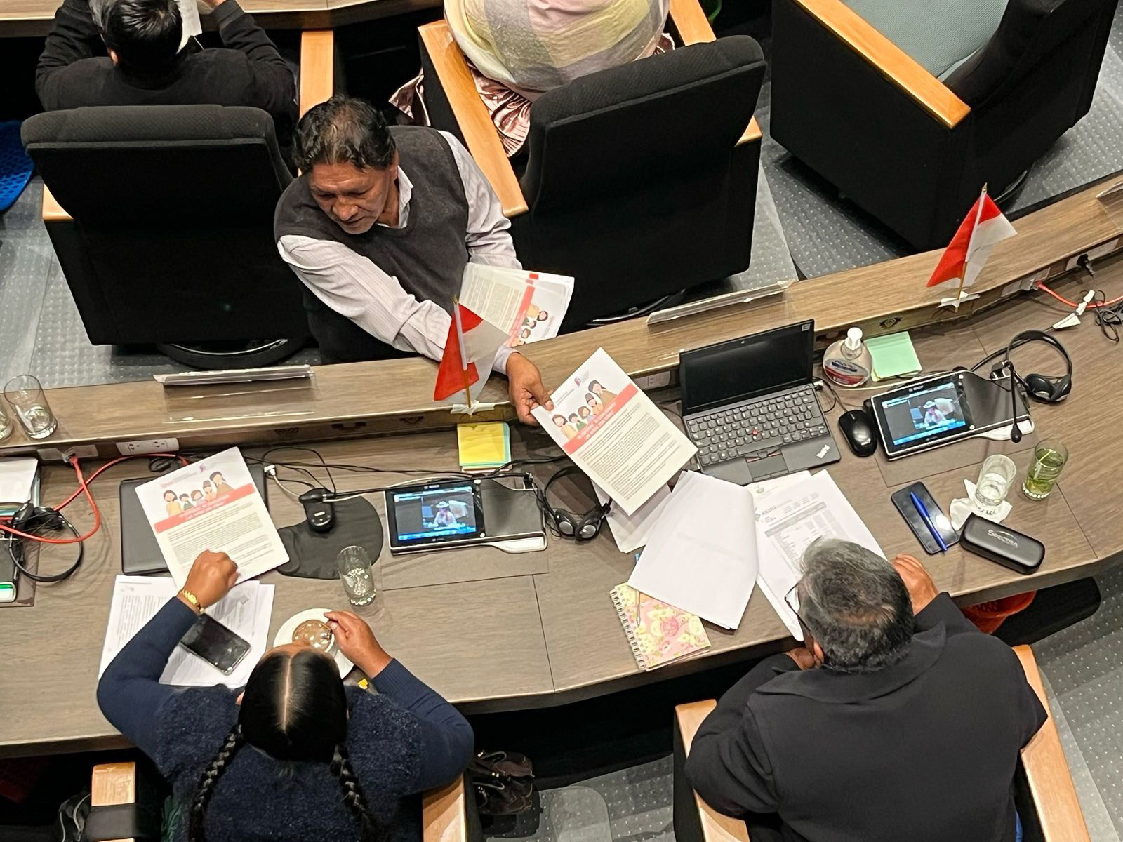 Legislators review information pamphlet prepared by Ipas Bolivia A group of people sit at curved desks, working with documents, laptops, and tablets. Small Peruvian flags are on the desks. One person hands papers to another. The scene appears to be a meeting or conference.