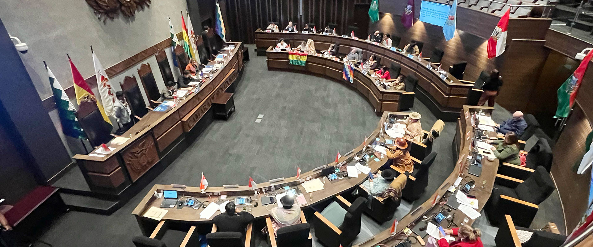 A large, curved conference table with people seated around it in a formal meeting room, each place marked with national flags. Several people are using laptops and microphones, and a presentation is displayed on a screen.
