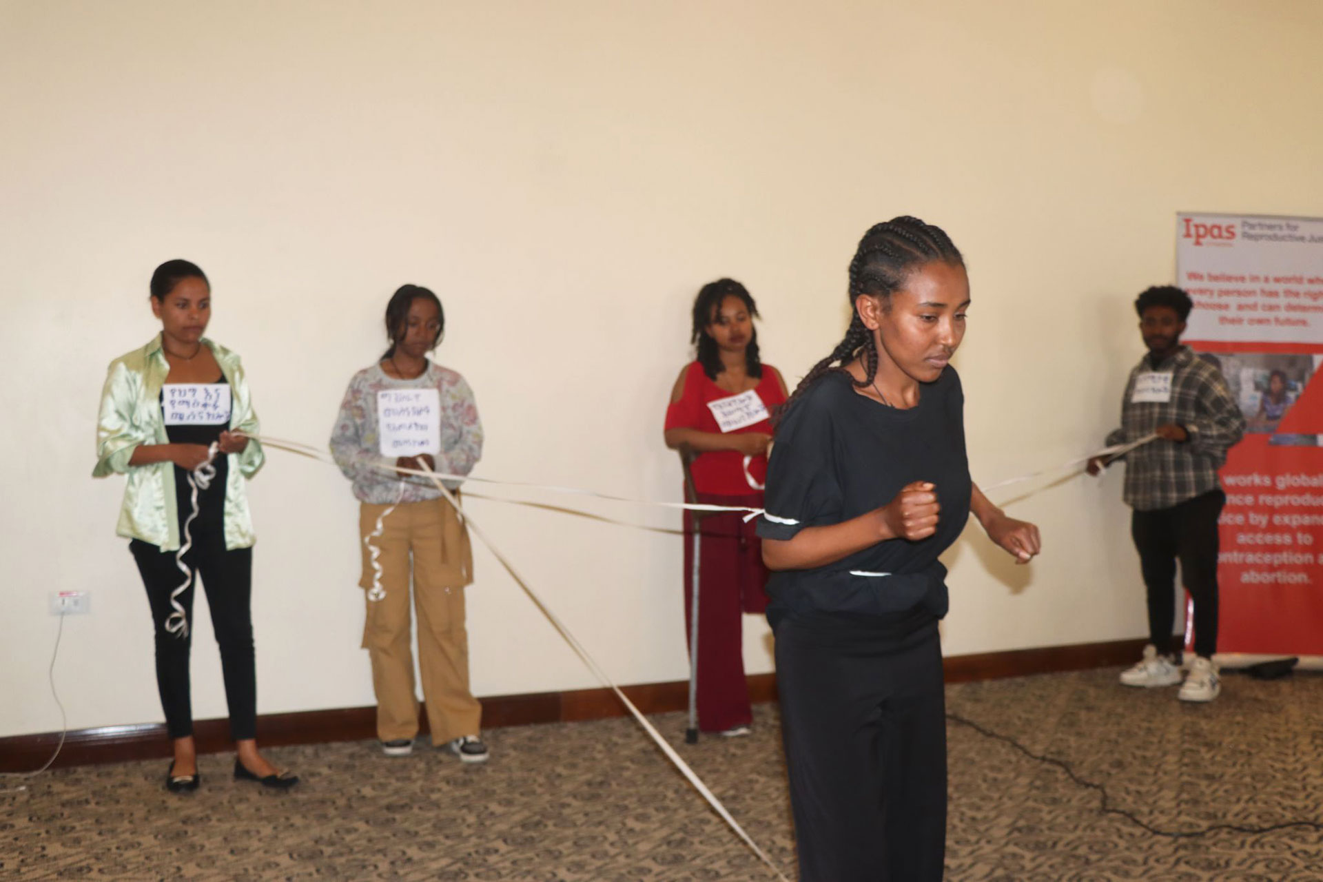 International Disability Day workshop Four people standing in a row hold onto strips of paper with signs on their chests, while a fifth person in front appears to be moving forward, pulling against the strips. A poster is visible on the right wall.