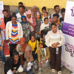 A group of men and women of various ages pose and smile together outdoors in front of two banners, one reading “Make Way,” against a yellow wall and stone pavement. Some are standing while others are kneeling or sitting.