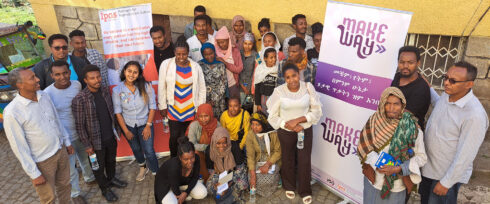 A group of men and women of various ages pose and smile together outdoors in front of two banners, one reading “Make Way,” against a yellow wall and stone pavement. Some are standing while others are kneeling or sitting.