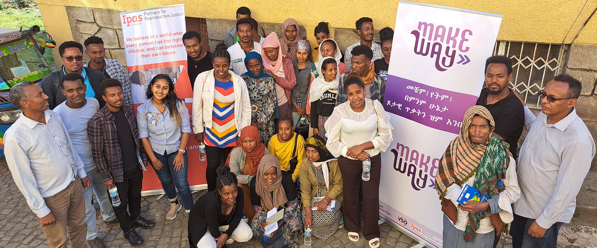 A group of men and women of various ages pose and smile together outdoors in front of two banners, one reading “Make Way,” against a yellow wall and stone pavement. Some are standing while others are kneeling or sitting.