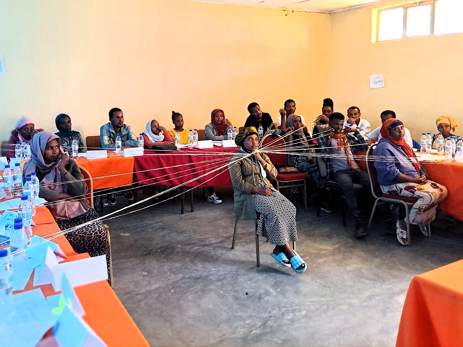 At a VCAT training for healthcare professionals in Dere Birhan A group of people sits in a classroom arranged in a U-shape. One woman sits in the center holding strings connected to others around her. Name cards and bottled water are on the tables. The room has orange tablecloths and a pale wall.