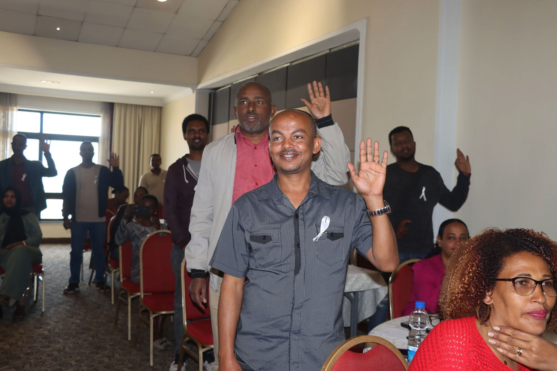 Community workshop, men pledge against GBV A group of people in a conference room stand and raise their right hands, some smiling at the camera, while others sit at round tables with red chairs. The atmosphere appears formal but friendly.