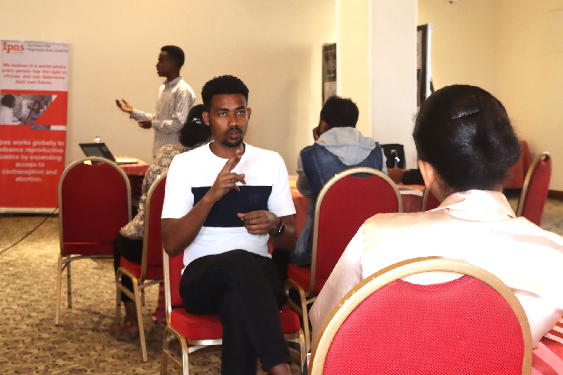 Community workshop, men pledge against GBV A group of people in a conference room stand and raise their right hands, some smiling at the camera, while others sit at round tables with red chairs. The atmosphere appears formal but friendly.