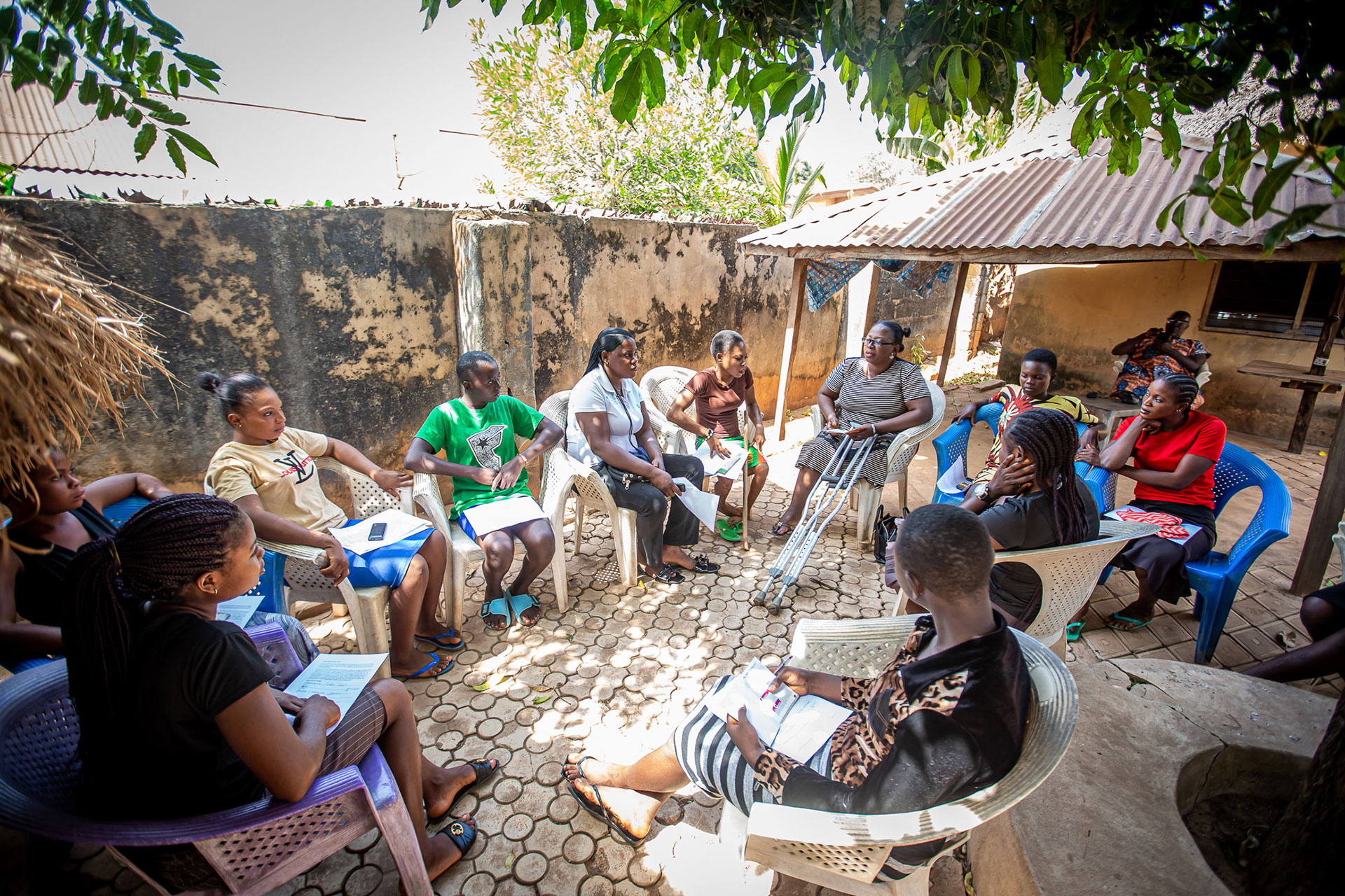 Peer-to-peer educators reached out to community groups. A group of women sit in a circle outdoors on plastic chairs, engaged in a discussion. They hold notebooks and pens, shaded by trees and a corrugated metal roof, near a rustic building with a weathered wall.