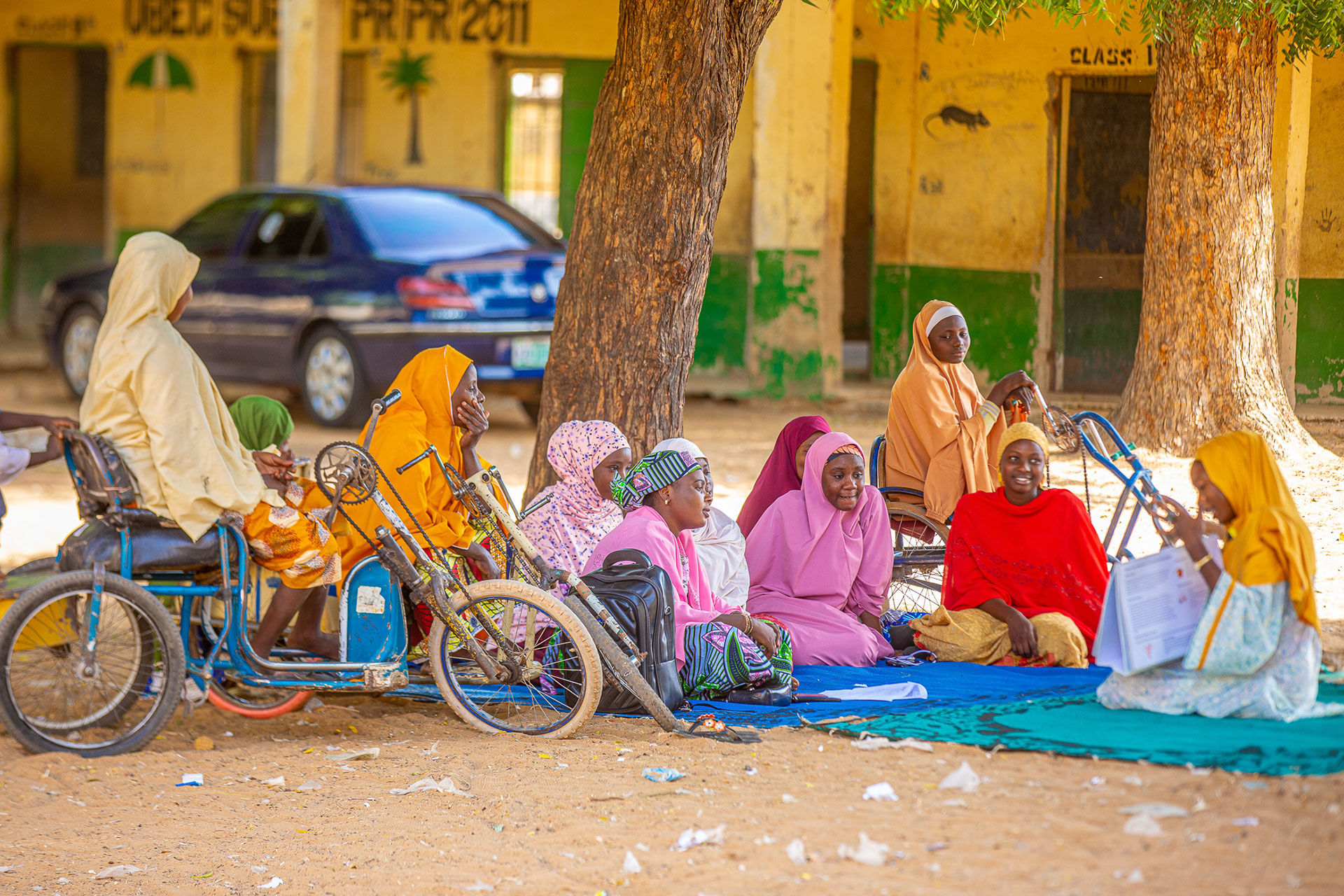 Peer-to-peer educators reached out to community groups. A group of women and girls in colorful hijabs sit on mats under a tree, engaging in a discussion or lesson outdoors. Bicycles are nearby, and a building with faded paint is visible in the background.
