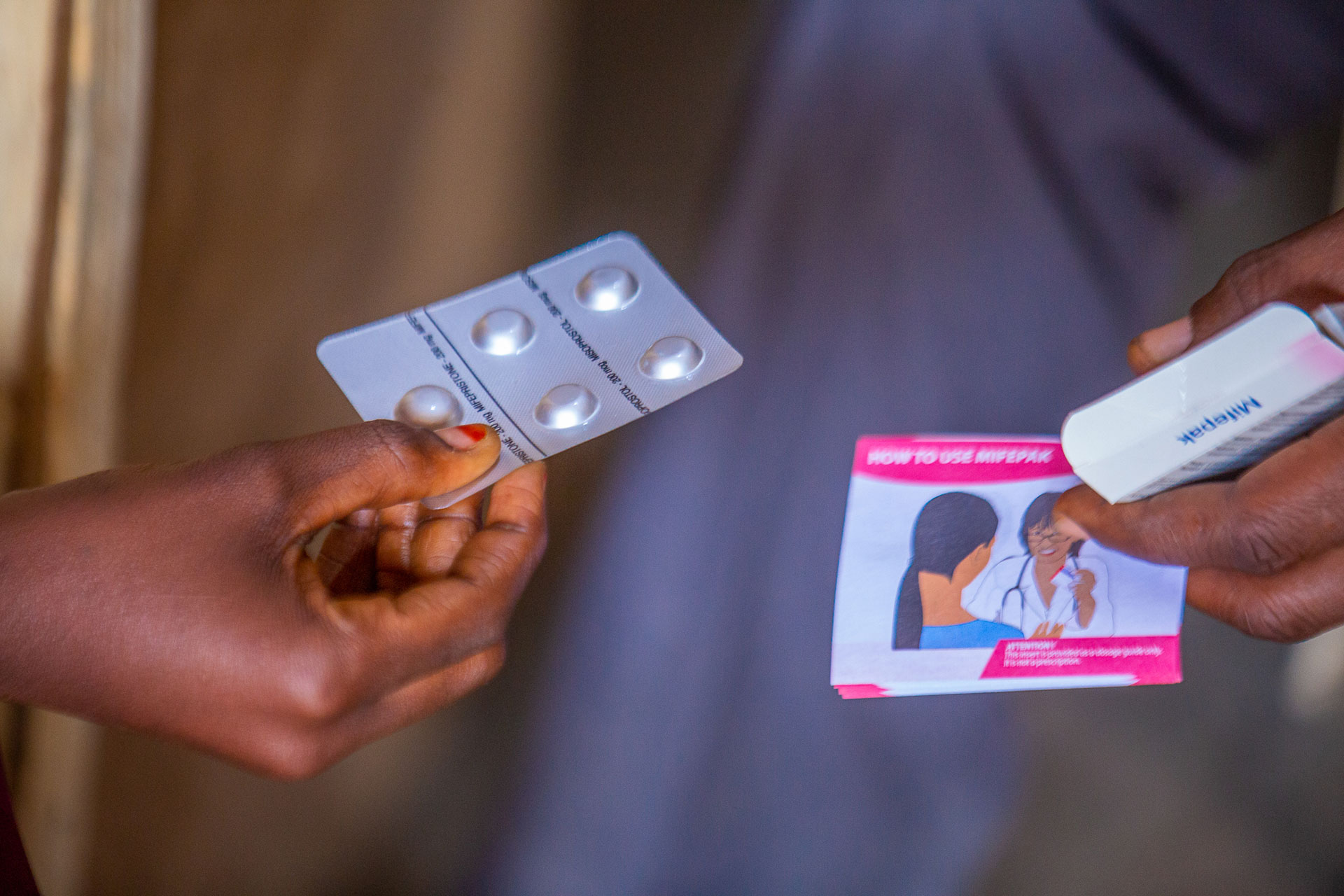 Two hands exchange a blister pack of four tablets and an instruction leaflet that shows a drawing of a woman and guidance on how to use medication. Two hands exchange a blister pack of four tablets and an instruction leaflet that shows a drawing of a woman and guidance on how to use medication.