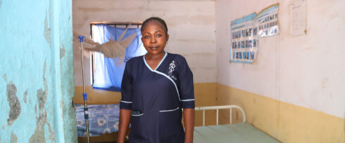 A woman in a navy nurse uniform stands in a simple clinic room with yellow and white walls, a bed, and medical posters. Light comes through a window covered by a torn blue curtain and mosquito net.