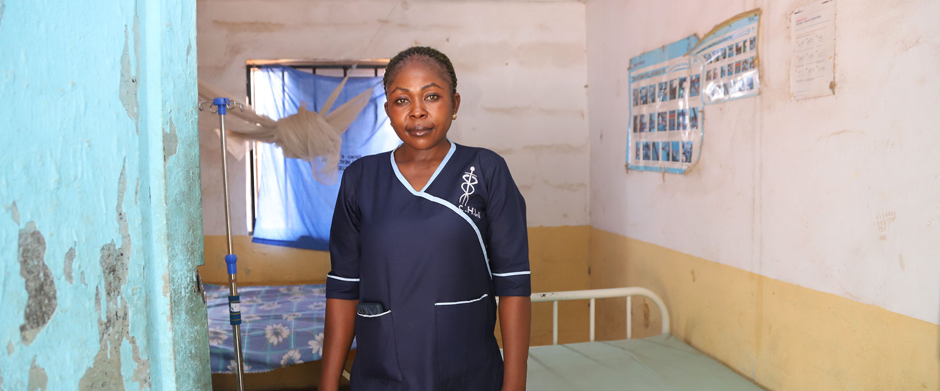 A woman in a navy nurse uniform stands in a simple clinic room with yellow and white walls, a bed, and medical posters. Light comes through a window covered by a torn blue curtain and mosquito net.