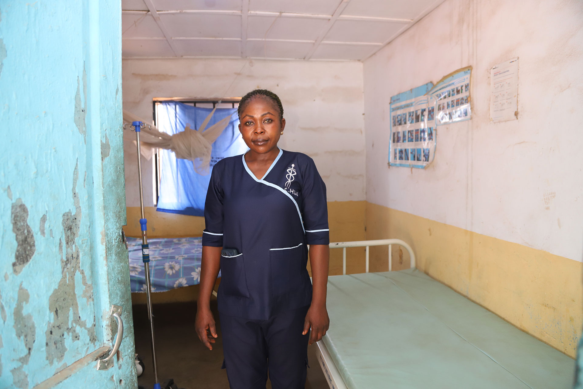 Suzan, one of the Ipas trained healthcare workers in Gboko South, Benue State. A woman in a navy blue nurse uniform stands in a small, simple clinic room with a single bed, IV stand, and medical posters on the wall. Light filters in through a blue-curtained window.