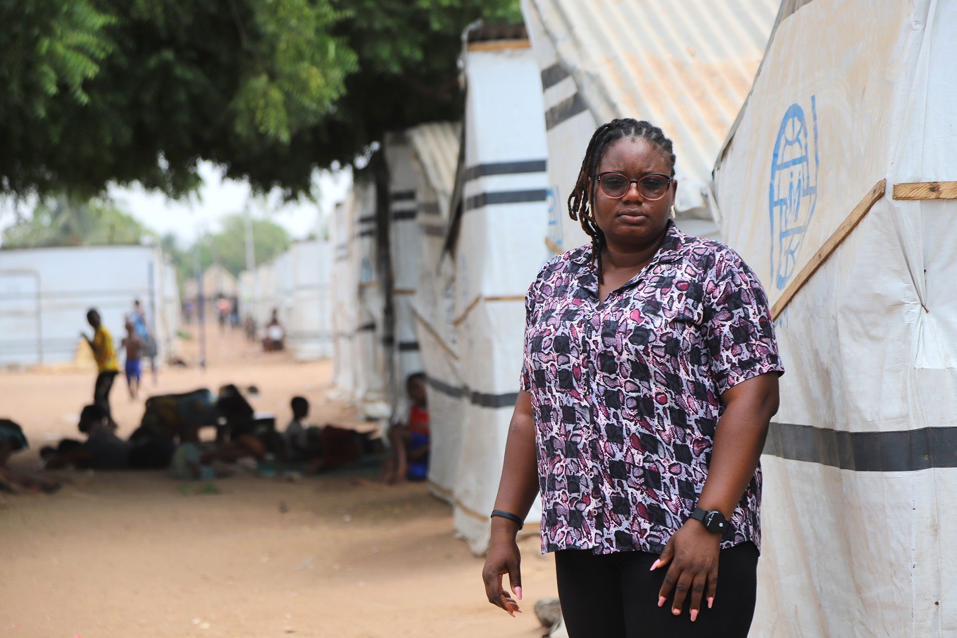 Deborah at one of the Internally Displaced Camps in Makurdi, Benue State. A woman wearing glasses and a patterned shirt stands in front of white tents in an outdoor camp area, with people sitting on the ground in the background under a tree.