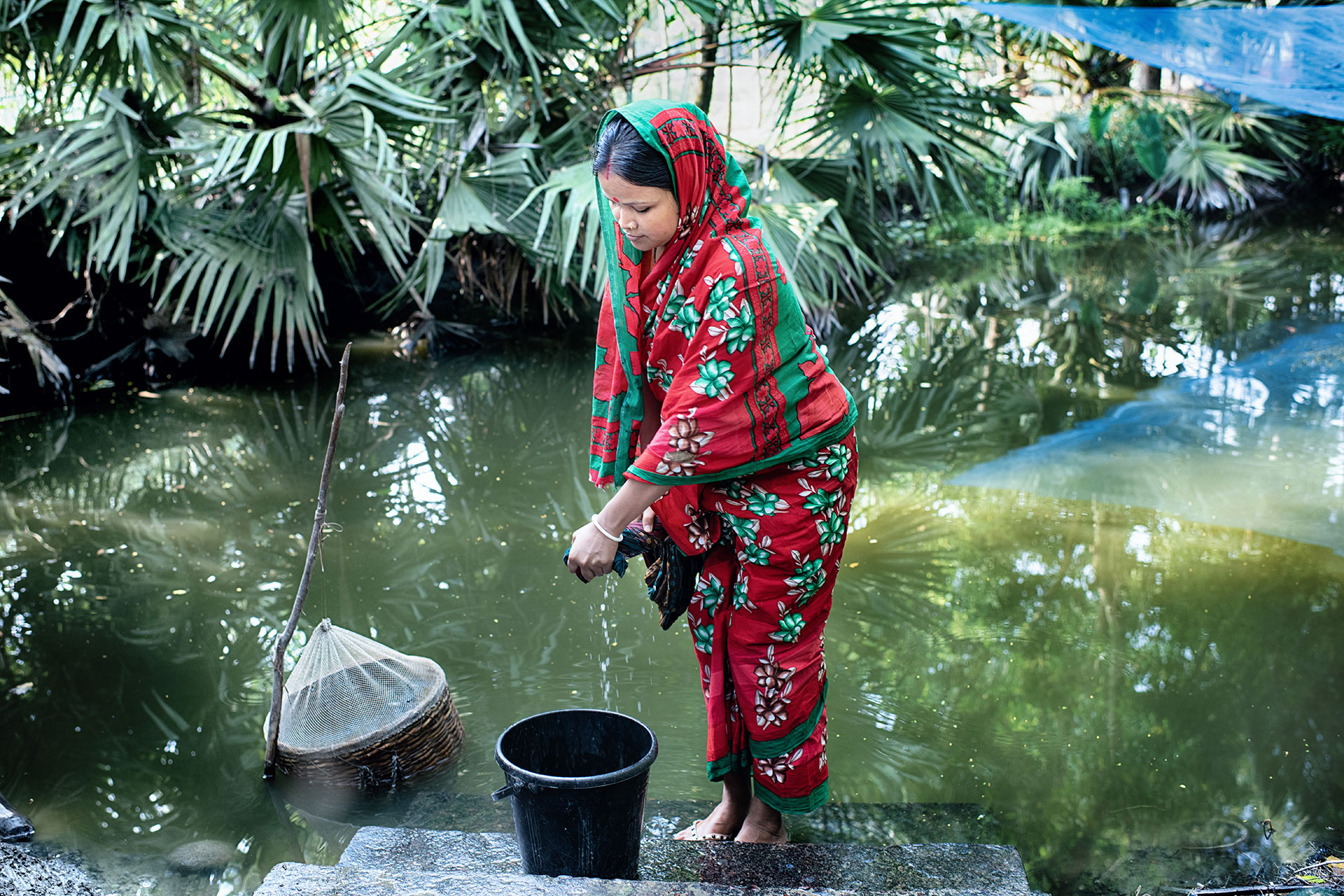 Trishna Bala Mondol, Shamnagar, Shatkhira, Bangladesh A woman in a red and green patterned sari collects water with a bucket from a pond, standing barefoot on stone steps surrounded by lush green foliage.