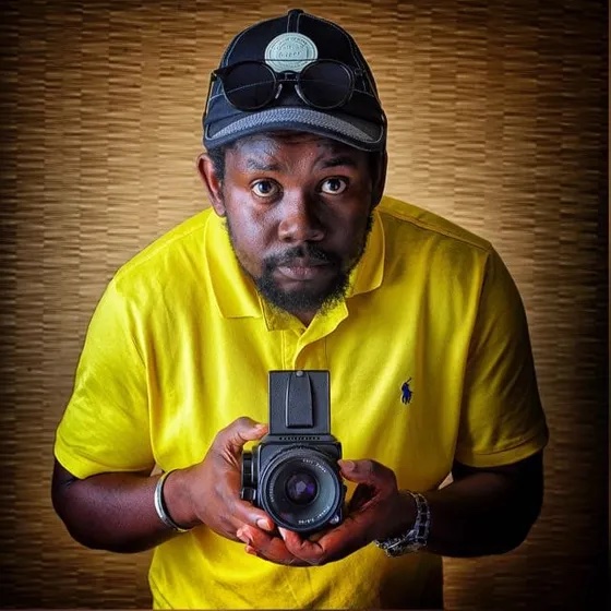 Photographer Amilton Neves A man in a yellow polo shirt and black cap, with sunglasses resting on his cap, looks at the camera while holding a vintage film camera against a textured brown background.