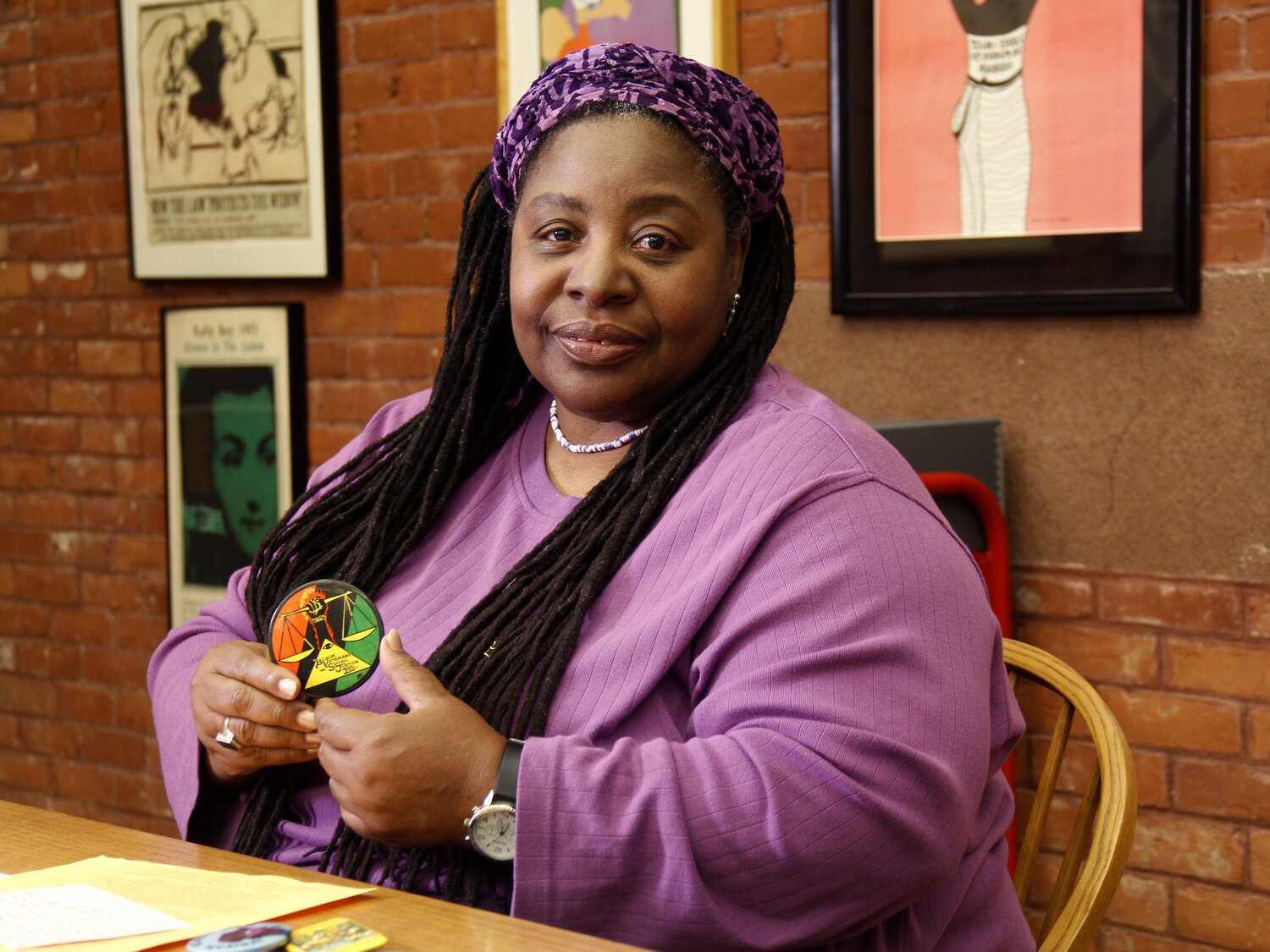 Loretta J. Ross Loretta J. Ross has long braids, wearing a purple outfit and headscarf, sits at a desk holding a colorful circular object. She is in a room with brick walls and framed artwork behind her.
