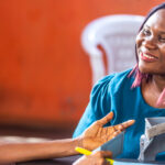 A healthcare worker speaks with a smiling woman seated at a table, holding a blood pressure cuff, in a brightly lit room with orange walls and a white plastic chair in the background.