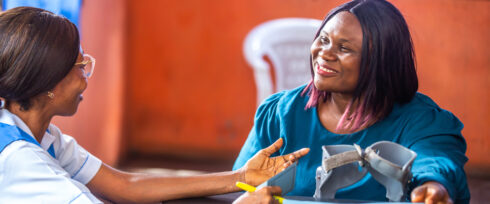 A healthcare worker speaks with a smiling woman seated at a table, holding a blood pressure cuff, in a brightly lit room with orange walls and a white plastic chair in the background.