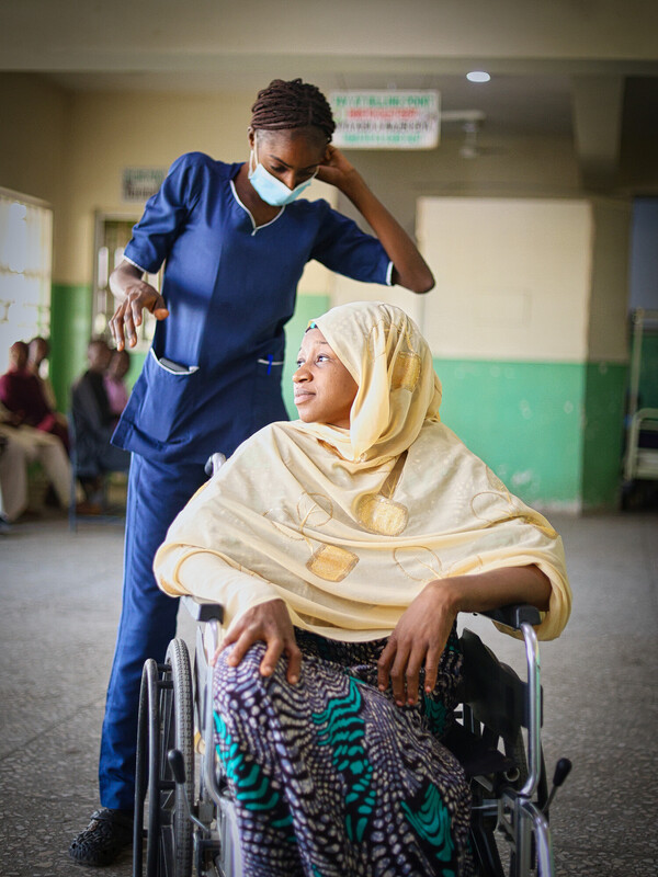 A nurse in blue scrubs and a face mask stands beside a woman in a patterned skirt and light hijab, who is sitting in a wheelchair, looking up at the nurse in a brightly lit, green-walled room. A nurse in blue scrubs and a face mask stands beside a woman in a patterned skirt and light hijab, who is sitting in a wheelchair, looking up at the nurse in a brightly lit, green-walled room.