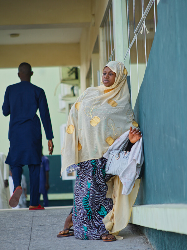 A woman in a patterned dress and light-colored hijab leans against a green and cream building, holding a bag, while a man walks away in the background. A woman in a patterned dress and light-colored hijab leans against a green and cream building, holding a bag, while a man walks away in the background.