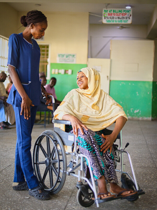 A nurse in blue scrubs smiles and talks with a woman in a wheelchair wearing a yellow hijab, patterned pants, and a beige top, inside a hospital waiting area with green and beige walls. A nurse in blue scrubs smiles and talks with a woman in a wheelchair wearing a yellow hijab, patterned pants, and a beige top, inside a hospital waiting area with green and beige walls.