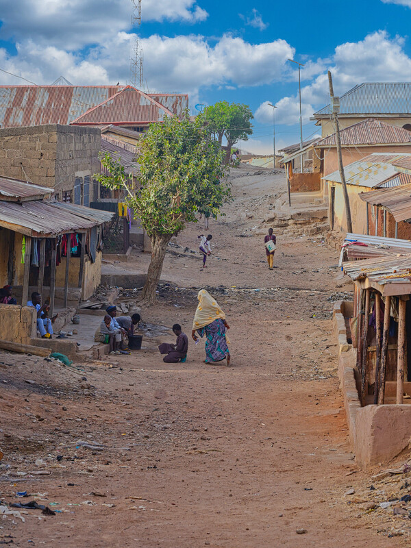 A dirt road runs through a rustic village with tin-roofed houses on both sides. Several people walk or sit along the street under a bright blue sky with scattered clouds. A dirt road runs through a rustic village with tin-roofed houses on both sides. Several people walk or sit along the street under a bright blue sky with scattered clouds.