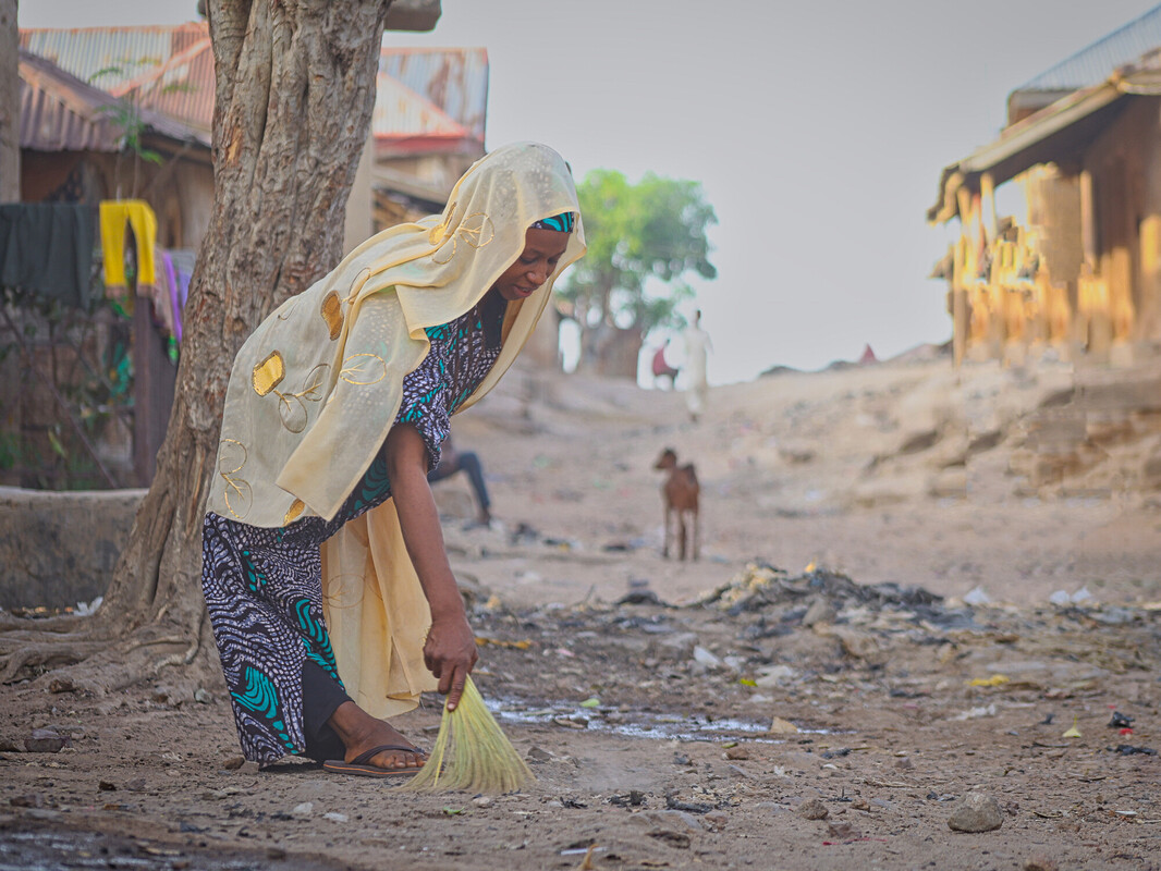 A woman in a patterned dress and light headscarf sweeps the dusty ground near a tree in a rural village setting, with houses on either side and a dog in the background. A woman in a patterned dress and light headscarf sweeps the dusty ground near a tree in a rural village setting, with houses on either side and a dog in the background.