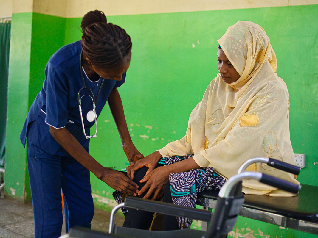 A healthcare worker in blue scrubs examines a woman's knee as she sits in a wheelchair, both are indoors against a green and yellow wall. The woman wears a light yellow headscarf and patterned clothing. A healthcare worker in blue scrubs examines a woman's knee as she sits in a wheelchair, both are indoors against a green and yellow wall. The woman wears a light yellow headscarf and patterned clothing.