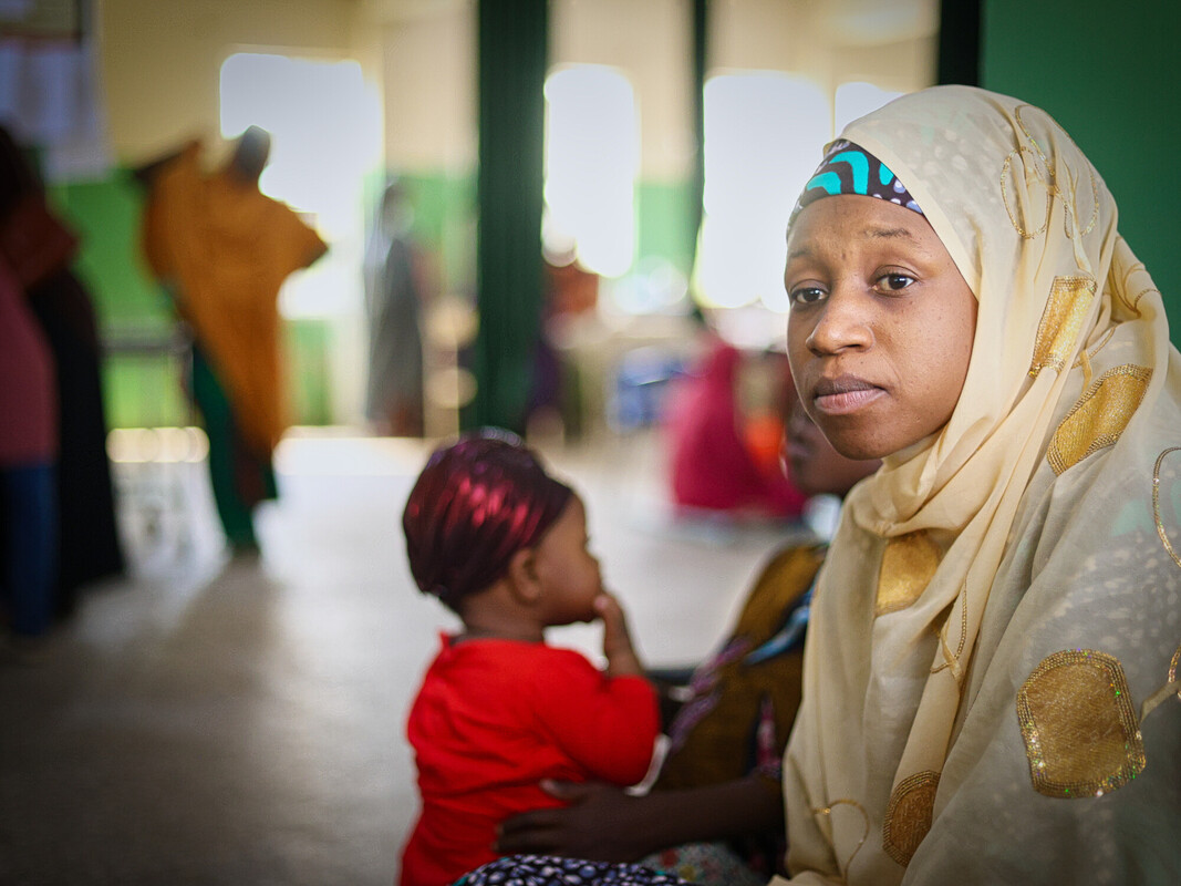 A woman in a cream hijab with gold patterns sits indoors, looking at the camera. A young child in a red outfit sits beside her. The background is softly blurred, showing other people and green walls. A woman in a cream hijab with gold patterns sits indoors, looking at the camera. A young child in a red outfit sits beside her. The background is softly blurred, showing other people and green walls.