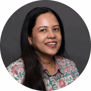 Portrait of Susmita Das. She is a woman with long dark hair, smiling at the camera. She is wearing a floral patterned shirt and is posed in front of a plain dark background inside a circular frame.