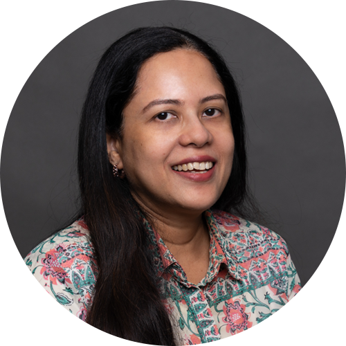 A woman with long dark hair smiles at the camera. She is wearing a floral patterned shirt and is posed in front of a plain dark background inside a circular frame. A woman with long dark hair smiles at the camera. She is wearing a floral patterned shirt and is posed in front of a plain dark background inside a circular frame.