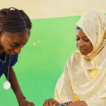 A nurse in blue scrubs checks the blood pressure of a woman wearing a light yellow hijab and patterned dress, seated next to a walker, against a green and cream wall.