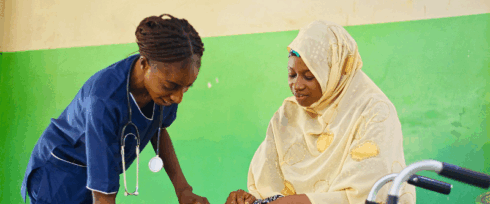 A nurse in blue scrubs checks the blood pressure of a woman wearing a light yellow hijab and patterned dress, seated next to a walker, against a green and cream wall.