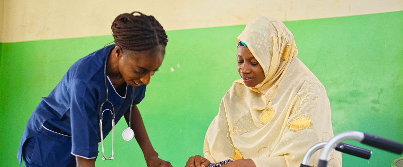A nurse in blue scrubs checks the blood pressure of a woman wearing a light yellow hijab and patterned dress, seated next to a walker, against a green and cream wall.