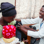 A healthcare worker in a white coat smiles while measuring a woman’s blood pressure; the woman holds a baby dressed in a red and white hat on her lap in a small clinic room.