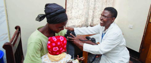 A healthcare worker in a white coat smiles while measuring a woman’s blood pressure; the woman holds a baby dressed in a red and white hat on her lap in a small clinic room.